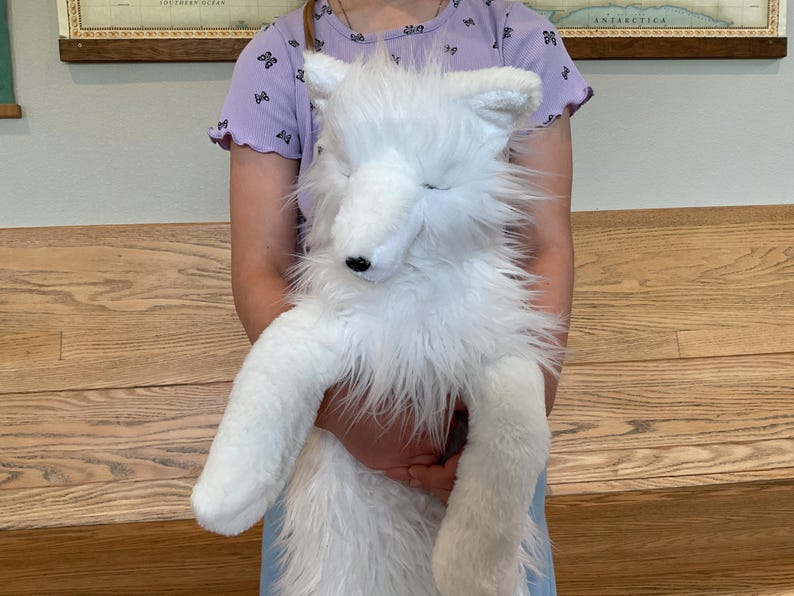 May include: A large, fluffy white plush fox toy with a black nose and closed eyes. The toy has long, soft fur and is being held by a person. The fox has long legs and a tail. The background includes a wooden bench and a map.