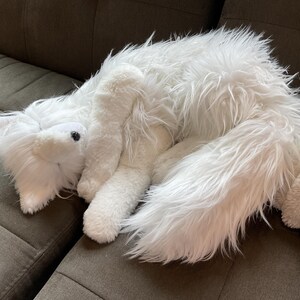 May include: A white fox soft toy with long, fluffy fur rests on a brown sofa. The toy fox has a pointed snout, black eyes, and a long, bushy tail. The plush toy appears soft and cuddly.