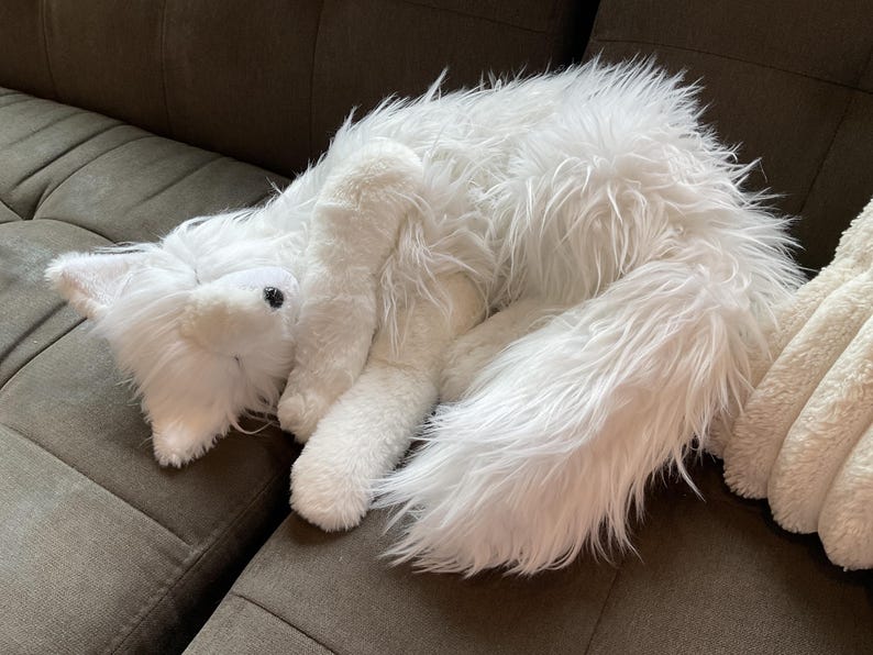 May include: A white fox soft toy with long, fluffy fur rests on a brown sofa. The toy fox has a pointed snout, black eyes, and a long, bushy tail. The plush toy appears soft and cuddly.