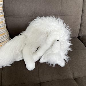 May include: A white, fluffy fox toy is lying on a brown sofa. The toy is curled up, with its head and tail visible. A decorative cushion with yellow, grey, and white stripes is in the background.