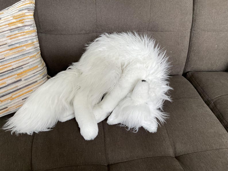 May include: A white, fluffy fox toy is lying on a brown sofa. The toy is curled up, with its head and tail visible. A decorative cushion with yellow, grey, and white stripes is in the background.