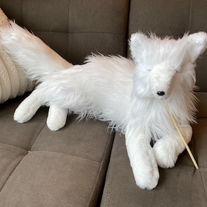 May include: A white plush fox toy with a long, fluffy tail and closed eyes. The fox is lying on a brown sofa, with a beige cushion in the background. A wooden stick is in its mouth.
