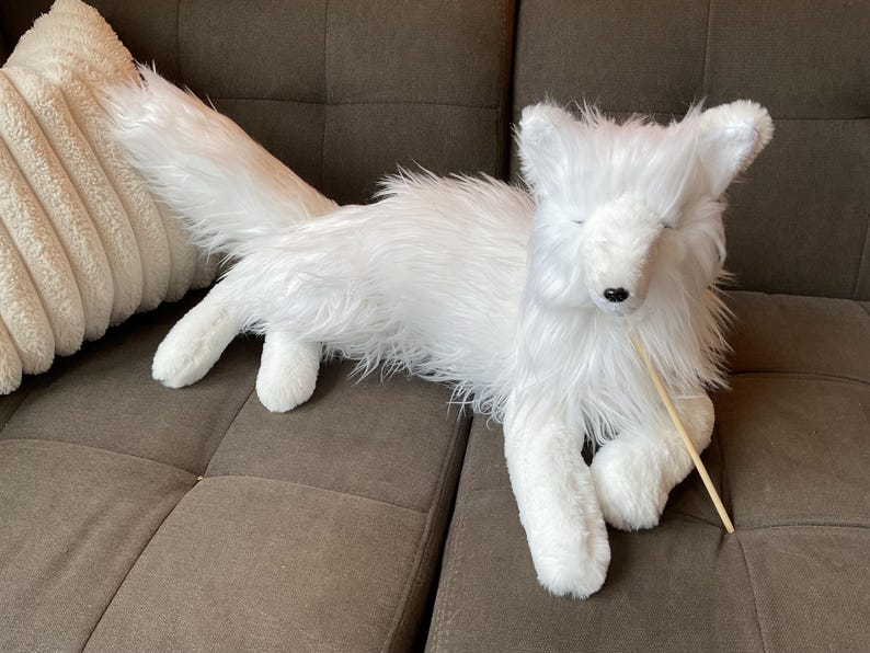May include: A white plush fox toy with a long, fluffy tail and closed eyes. The fox is lying on a brown sofa, with a beige cushion in the background. A wooden stick is in its mouth.