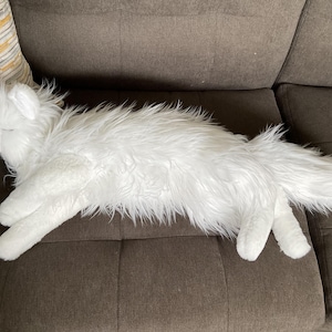 May include: A white plush wolf toy with long, shaggy fur, resting on a brown sofa. The toy has a detailed head, paws, and a fluffy tail. The wolf is positioned horizontally, appearing relaxed.