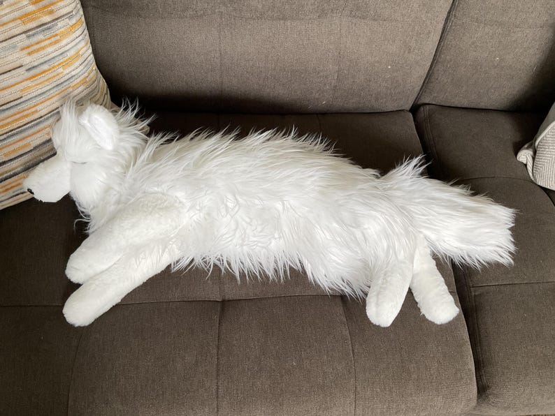 May include: A white plush wolf toy with long, shaggy fur, resting on a brown sofa. The toy has a detailed head, paws, and a fluffy tail. The wolf is positioned horizontally, appearing relaxed.