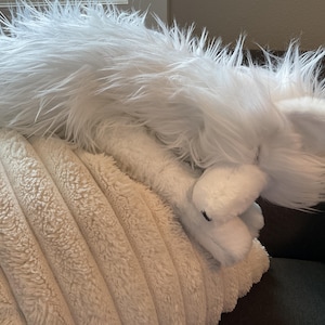 May include: A white, fluffy toy fox with long fur, lying on a cream-coloured, textured pillow. The fox has visible ears and a soft appearance, suggesting a children's toy or decorative item.