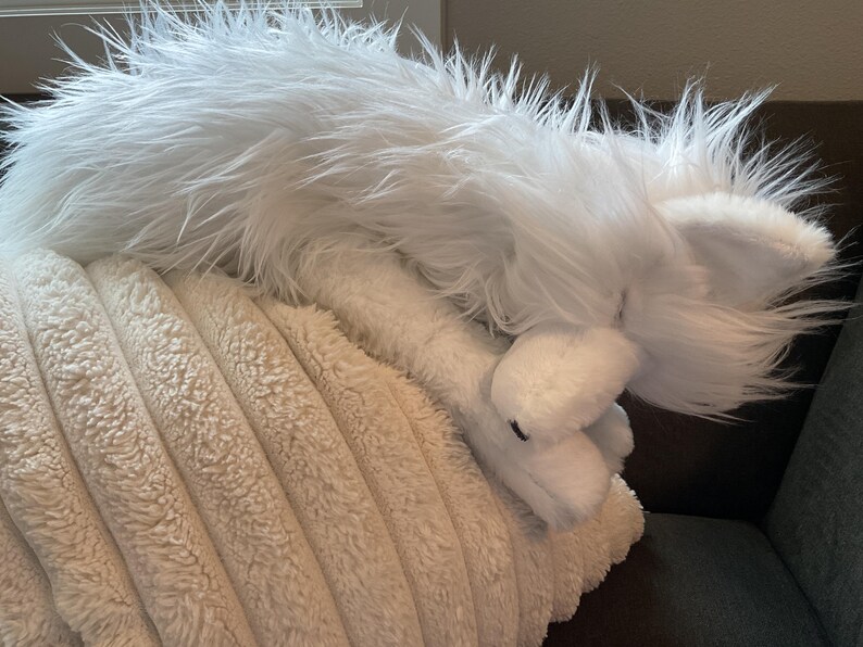 May include: A white, fluffy toy fox with long fur, lying on a cream-coloured, textured pillow. The fox has visible ears and a soft appearance, suggesting a children's toy or decorative item.