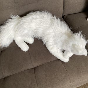 May include: A white, furry animal-shaped cushion lies on a brown sofa. The cushion has a long, fluffy tail and is positioned as if lying down. The sofa has two decorative cushions.