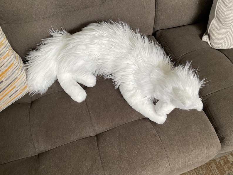 May include: A white, furry animal-shaped cushion lies on a brown sofa. The cushion has a long, fluffy tail and is positioned as if lying down. The sofa has two decorative cushions.