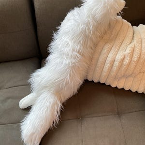 May include: A white, fluffy cat-shaped cushion with long fur. The cushion is on a brown corduroy pillow and a brown sofa. The cat pillow has a long tail and pointed ears.