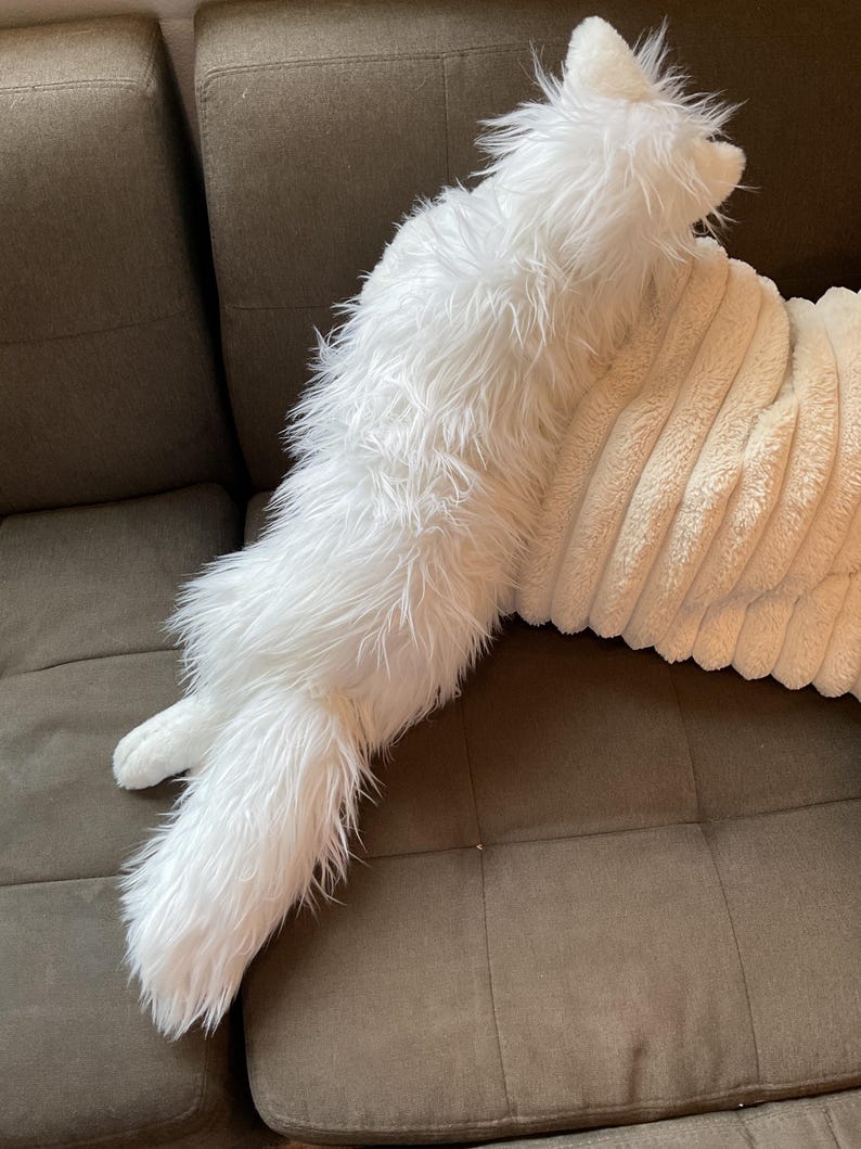 May include: A white, fluffy cat-shaped cushion with long fur. The cushion is on a brown corduroy pillow and a brown sofa. The cat pillow has a long tail and pointed ears.