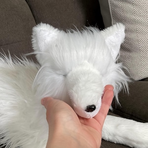 May include: A white plush fox toy with a black nose and closed eyes. The toy has long, fluffy fur and is being held by a hand. The fox is lying on a dark brown surface.