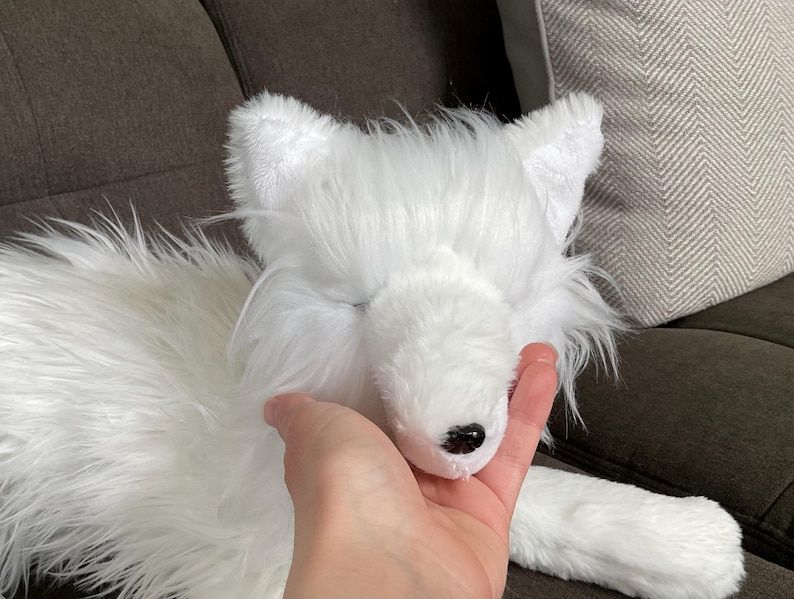 May include: A white plush fox toy with a black nose and closed eyes. The toy has long, fluffy fur and is being held by a hand. The fox is lying on a dark brown surface.
