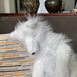 May include: A fluffy white toy dog with long, shaggy fur is resting on a striped cushion. The dog has a black nose and is lying on a sofa. The cushion has a pattern of yellow, grey, and white stripes.