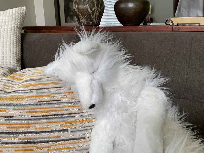 May include: A fluffy white toy dog with long, shaggy fur is resting on a striped cushion. The dog has a black nose and is lying on a sofa. The cushion has a pattern of yellow, grey, and white stripes.