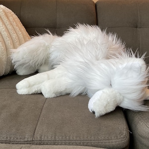 May include: A white, fluffy dog toy lies on a brown sofa. The toy has long, shaggy fur and is positioned near a cream-coloured, textured cushion. The dog toy appears to be in a relaxed position.