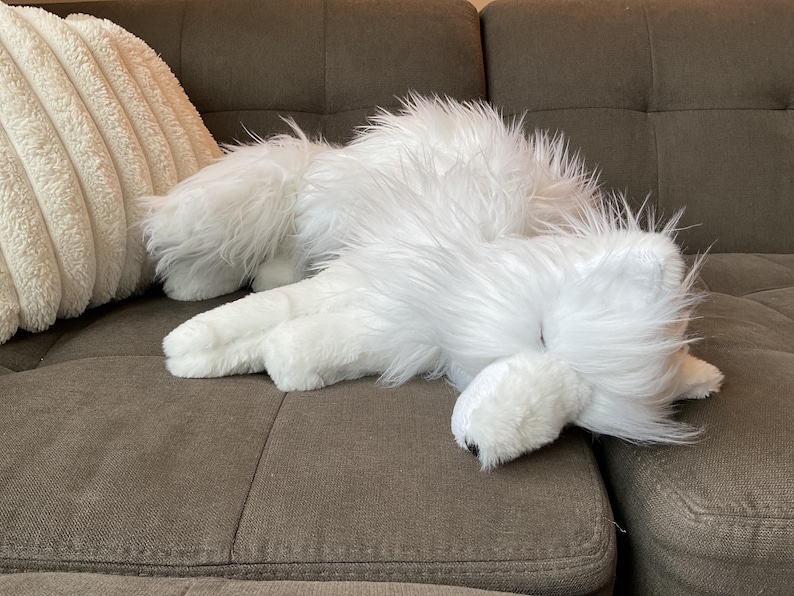 May include: A white, fluffy dog toy lies on a brown sofa. The toy has long, shaggy fur and is positioned near a cream-coloured, textured cushion. The dog toy appears to be in a relaxed position.
