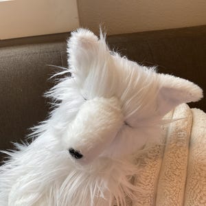May include: A white plush toy dog with closed eyes and a black nose. The dog has long, shaggy fur and is resting on a beige blanket. The ears are upright and the toy is positioned on a brown surface.