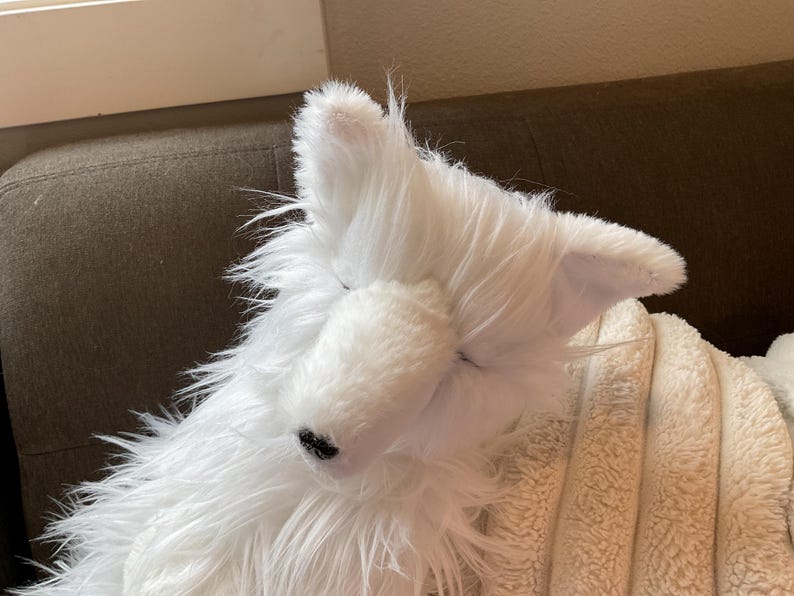 May include: A white plush toy dog with closed eyes and a black nose. The dog has long, shaggy fur and is resting on a beige blanket. The ears are upright and the toy is positioned on a brown surface.