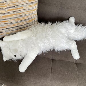 May include: A white, fluffy cat-shaped soft toy with long fur lies on a brown sofa. The toy has black button eyes and a long tail. A striped cushion with yellow, grey, and brown stripes is in the background.
