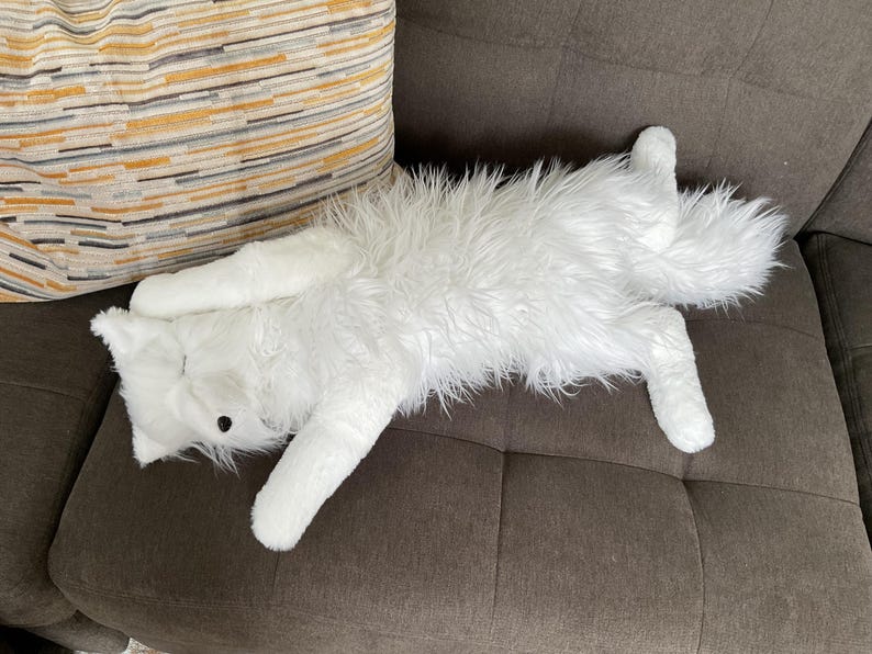 May include: A white, fluffy cat-shaped soft toy with long fur lies on a brown sofa. The toy has black button eyes and a long tail. A striped cushion with yellow, grey, and brown stripes is in the background.