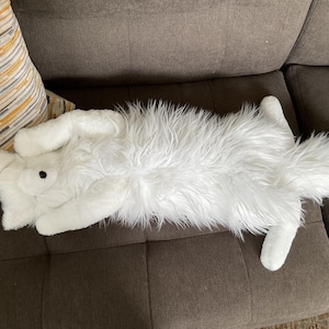 May include: A white, cat-shaped cushion with long, fluffy fur. The cushion has a black button eye and is lying on its back on a brown sofa. The cushion's tail is long and fluffy.