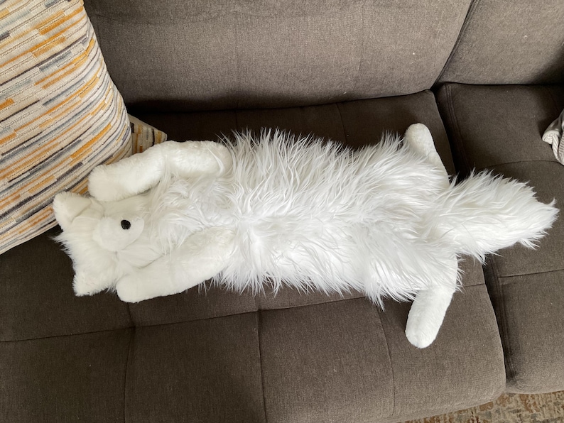 May include: A white, cat-shaped cushion with long, fluffy fur. The cushion has a black button eye and is lying on its back on a brown sofa. The cushion's tail is long and fluffy.