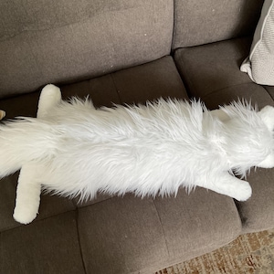 May include: A long, white, plush cat-shaped cushion with a fluffy fur texture. The cushion is lying on a brown sofa, showcasing its full body and detailed features. The cat cushion adds a touch of whimsy to home decor.