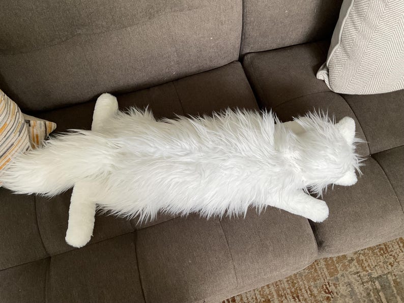 May include: A long, white, plush cat-shaped cushion with a fluffy fur texture. The cushion is lying on a brown sofa, showcasing its full body and detailed features. The cat cushion adds a touch of whimsy to home decor.