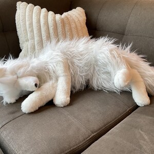 May include: A white, fluffy fox toy lies on a brown sofa, next to a cream-coloured, corduroy-textured cushion. The toy has a black nose and long, shaggy fur. Its legs are extended, and its head is resting, creating a relaxed pose.