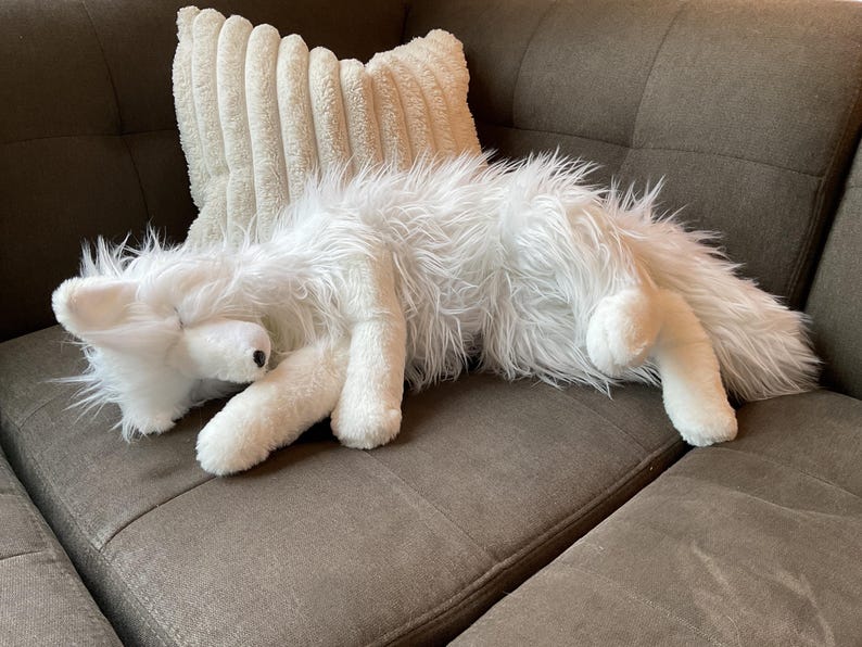 May include: A white, fluffy fox toy lies on a brown sofa, next to a cream-coloured, corduroy-textured cushion. The toy has a black nose and long, shaggy fur. Its legs are extended, and its head is resting, creating a relaxed pose.