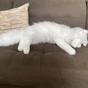 May include: A white, fluffy cat-shaped soft toy rests on a brown sofa. The toy is lying down, with its paws outstretched. A decorative cushion with horizontal stripes in yellow, grey, and white is visible in the background.