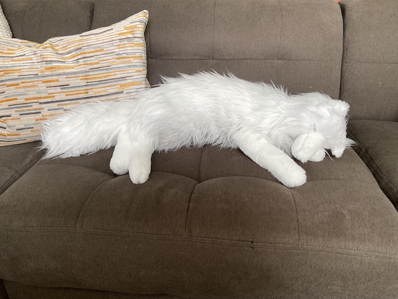 May include: A white, fluffy cat-shaped soft toy rests on a brown sofa. The toy is lying down, with its paws outstretched. A decorative cushion with horizontal stripes in yellow, grey, and white is visible in the background.