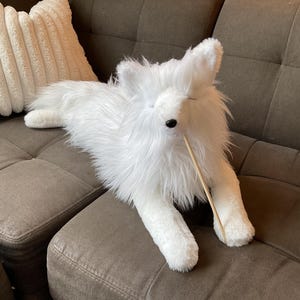 May include: A white plush fox toy with black eyes and a long, fluffy tail is lying on a brown sofa. The fox has a long, thin wooden stick in its mouth. A white, textured cushion is in the background.