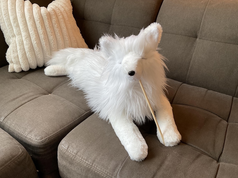 May include: A white plush fox toy with black eyes and a long, fluffy tail is lying on a brown sofa. The fox has a long, thin wooden stick in its mouth. A white, textured cushion is in the background.