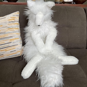 May include: A white plush fox toy with a shaggy, fluffy texture. The fox is seated with its eyes closed and paws crossed. It has a long, bushy tail. This is a decorative item.