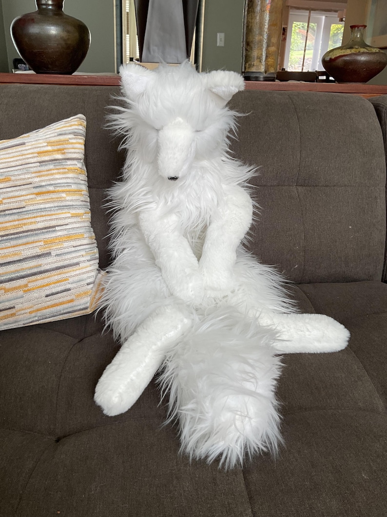 May include: A white plush fox toy with a shaggy, fluffy texture. The fox is seated with its eyes closed and paws crossed. It has a long, bushy tail. This is a decorative item.