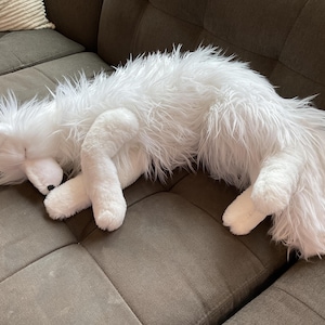May include: A white fox soft toy is curled up on a grey sofa. The plush animal has a shaggy, fluffy texture and is in a sleeping position. The fox's paws and face are visible, with a black nose.