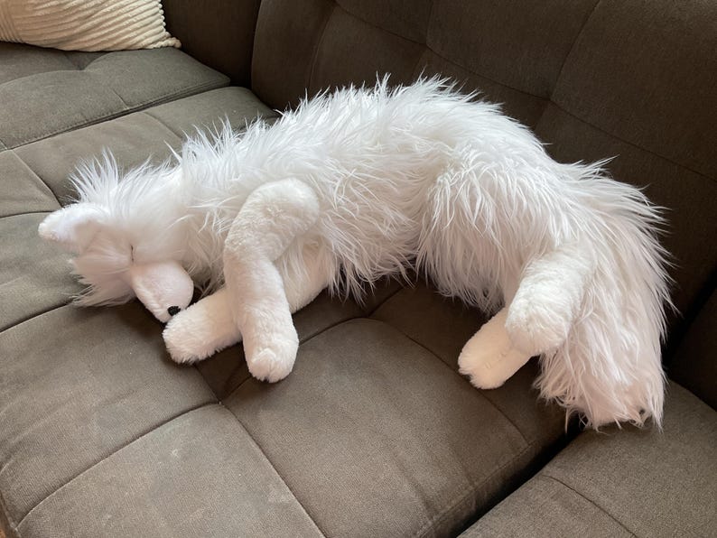 May include: A white fox soft toy is curled up on a grey sofa. The plush animal has a shaggy, fluffy texture and is in a sleeping position. The fox's paws and face are visible, with a black nose.