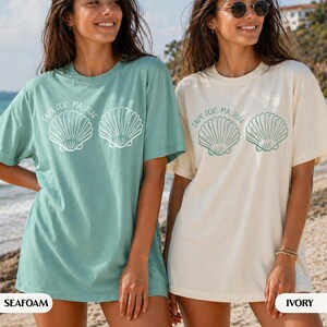 May include: Two short-sleeved t-shirts, one seafoam green and one ivory, each featuring a white seashell graphic and the text "CAPE COD, MA, 2026". The shirts are casual and suitable for beach or summer wear.