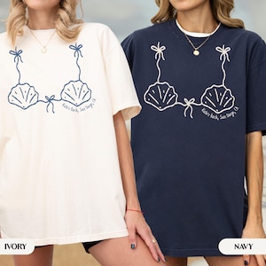 以下が含まれることがあります： アイボリーとネイビーの2枚のTシャツ。それぞれに白い貝殻ブラのグラフィックが描かれています。デザインにはリボンと「Kate's Bach, San Diego, CA」の文字が含まれています。Tシャツは半袖で、ゆったりとしたフィット感です。