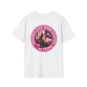 Puede incluir: Camiseta blanca con un gráfico circular rosa. El gráfico muestra tres gatos en una habitación y el texto "JUST DOING MY THING". Los gatos son negros, marrones y blancos.