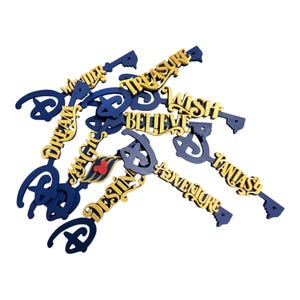 May include: A collection of dark blue key-shaped ornaments featuring gold lettering with the words Wonder, Treasure, Wish, Dream, Believe, Magic, Destiny, Adventure, and Fantasy. Each key includes a stylized D-shaped bow and a small red icon.