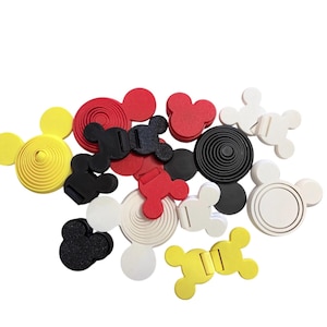 May include: A collection of small, multi-colored plastic accessories shaped like mouse heads. The items include yellow, red, black, and white pieces, some featuring concentric circular patterns or hinged mechanisms, scattered in a loose arrangement.
