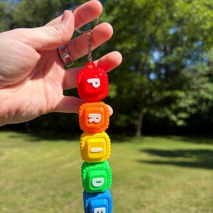 May include: A colorful keychain spelling "PRIDE" with red, orange, yellow, green, and blue blocks. Each block has a letter in white. The keychain is held up against a blurred green background.