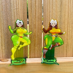 May include: Two acrylic figurines of a comic book character in a dynamic pose. One is in a green and yellow costume, the other in a brown and green outfit. Both have white hair and are on green bases, set against a wooden fence.