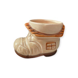 May include: A ceramic planter shaped like a boot with a house design. The boot is cream-colored with brown accents on the sole, the top rim, and the square window detail on the side. The surface has a glossy finish and molded textures.