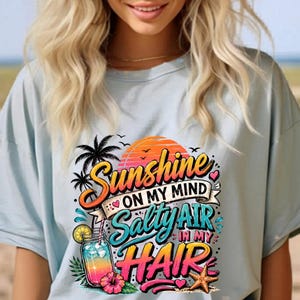 Könnte beinhalten: Hellblaues T-Shirt mit dem Text "Sunshine on my mind, Salty air in my hair". Die Grafik zeigt einen Sonnenuntergang, Palmen, ein Glas mit Getränk und einen Seestern. Das Shirt hat ein lässiges Strand-Design.