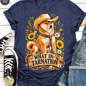 May include: A heather navy t-shirt featuring a graphic of a Shiba Inu dog wearing a tan cowboy hat and a patterned collar. The dog is surrounded by yellow sunflowers and white daisies. Below the dog is a wooden sign with the text WHAT IN TARNATION.