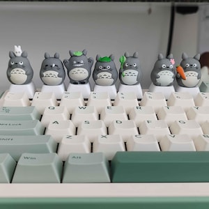 May include: A mechanical keyboard with white keycaps and a green accent. Adorning the top row are seven gray Totoro-themed keycap figures, each with unique accessories. The keyboard's design is clean and modern, with a focus on the custom keycaps.